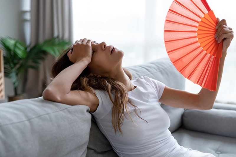 Why Is My AC Blowing Hot Air? Young woman using paper fan, suffering from hot summer weather or high temperature at home.