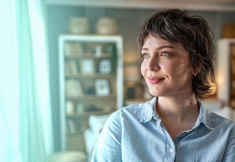 A portrait of a woman with short, wavy dark brown hair and light eyes, wearing a light blue denim shirt.