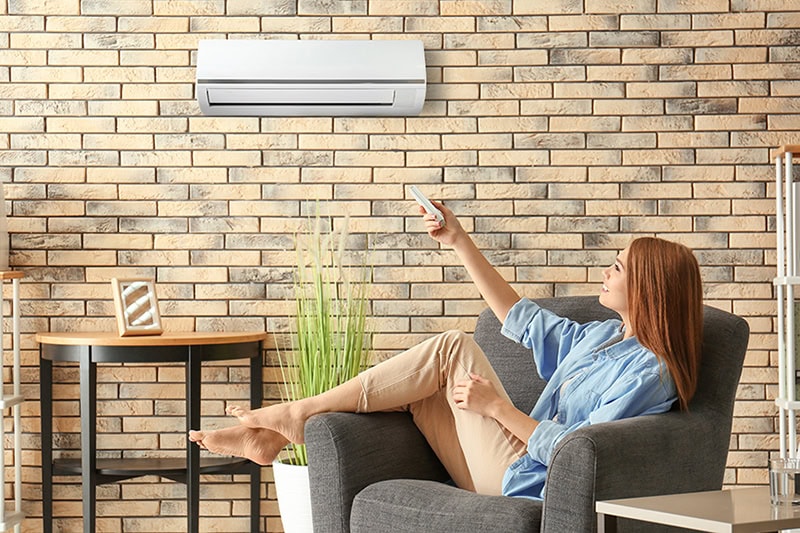 Hot and Cold Spots? Get Even Heating and Cooling in Your Home. Photo of a young woman switching on their air conditioner while sitting in a chair in her home.